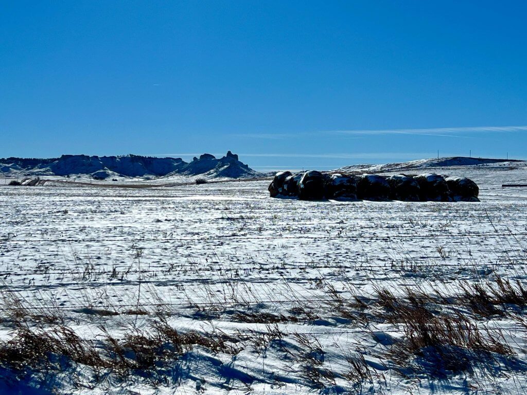 Pasture grasses poke through the snow covered fields. Hay stack with snow and high country buttes in the background. 